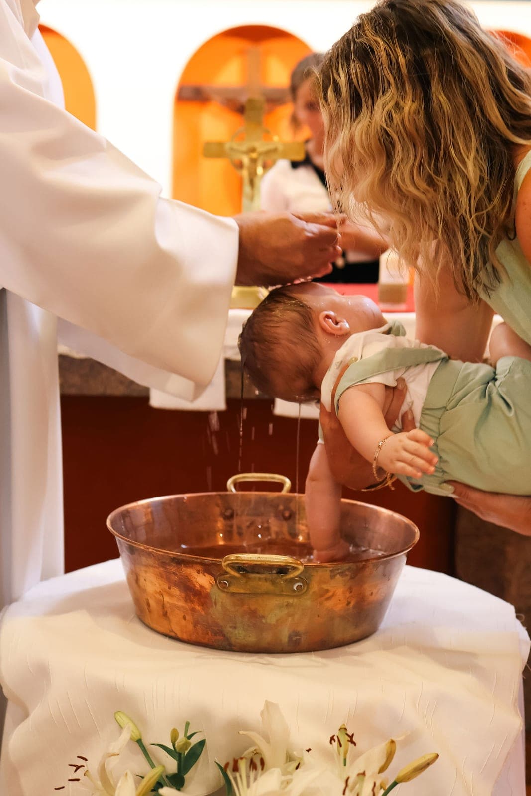 L’eau du baptême versée sur le front d’un bébé, au-dessus d’une vasque de cuivre.
