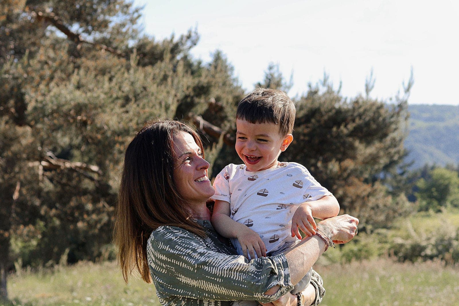 Une maman tient son petit garçon qui éclate de rire, dans un champ en fin de journée.