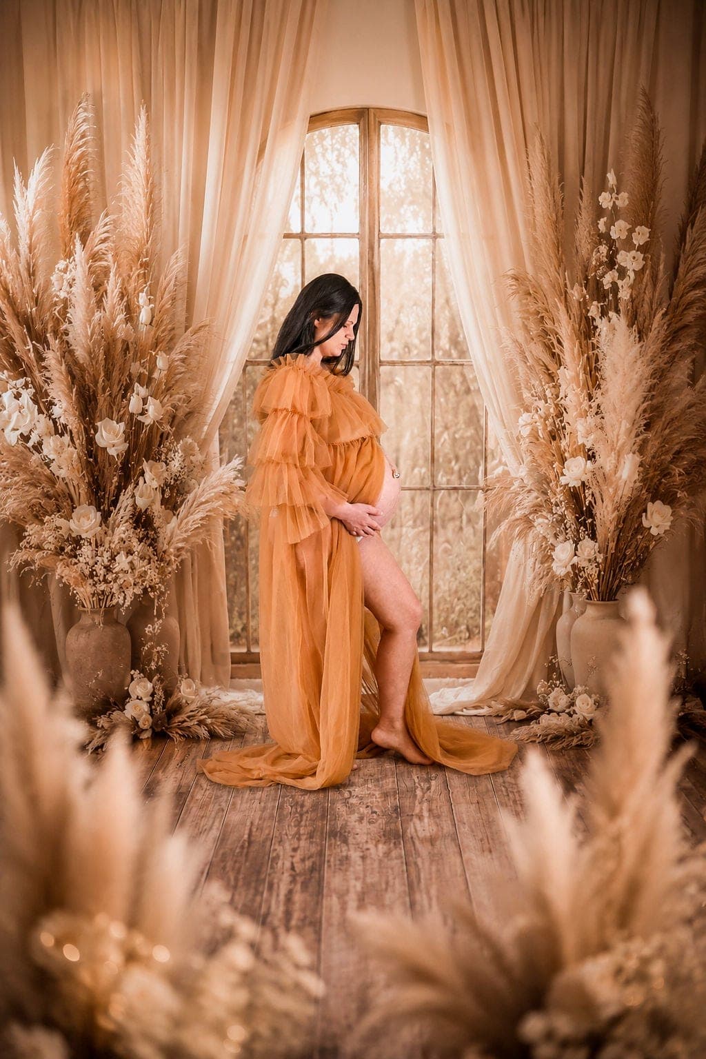 Future maman en robe safran, debout devant une fenêtre et des pampas, au studio.